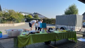 Hofverkauf im Freien mit verschiedenen Artikeln auf Tischen, einem Anhänger im Hintergrund und drei Personen, die während der sonnigen Veranstaltungen in der Nähe des Anhängers interagieren.