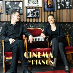 Zwei schwarz gekleidete Personen sitzen auf roten Theatersesseln, zwischen ihnen ein kleines Klavierklavier. An der Wand dahinter hängen gerahmte Fotos und Poster, die auf besondere Veranstaltungen hinweisen. Der Text auf dem Sitz lautet "CINEMA for PIANOS.