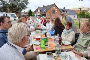 Eine Gruppe von Menschen sitzt an einem langen Tisch im Freien, der mit Speisen und Getränken gedeckt ist. Sie haben sich zu einer Mahlzeit in einem Wohnviertel versammelt und genießen eine der vielen Veranstaltungen, die die Gemeinschaft zusammenbringen.