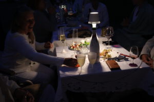 Menschen sitzen bei einer Abendveranstaltung um einen Tisch herum, mit Getränken, Essen und einer Lampe, die Licht spendet; eine Frau greift nach einem Glas und lächelt.