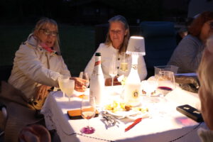 Vier Frauen sitzen abends im Freien an einem Tisch und genießen unter einer kleinen Lampe Getränke und Speisen - die perfekte Inspiration für intime Veranstaltungen oder die Planung zwangloser Events. Der Tisch ist mit Flaschen, Gläsern und Tellern gedeckt.