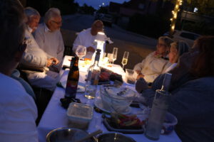 Eine Gruppe von Menschen sitzt in der Abenddämmerung im Freien an einem Tisch und teilt unter Lichterketten Getränke und Essen. Flaschen, Schüsseln und Essensbehälter stehen auf dem Tisch und fangen die warme Stimmung ein, die man oft bei Veranstaltungen in Deutschland sieht.